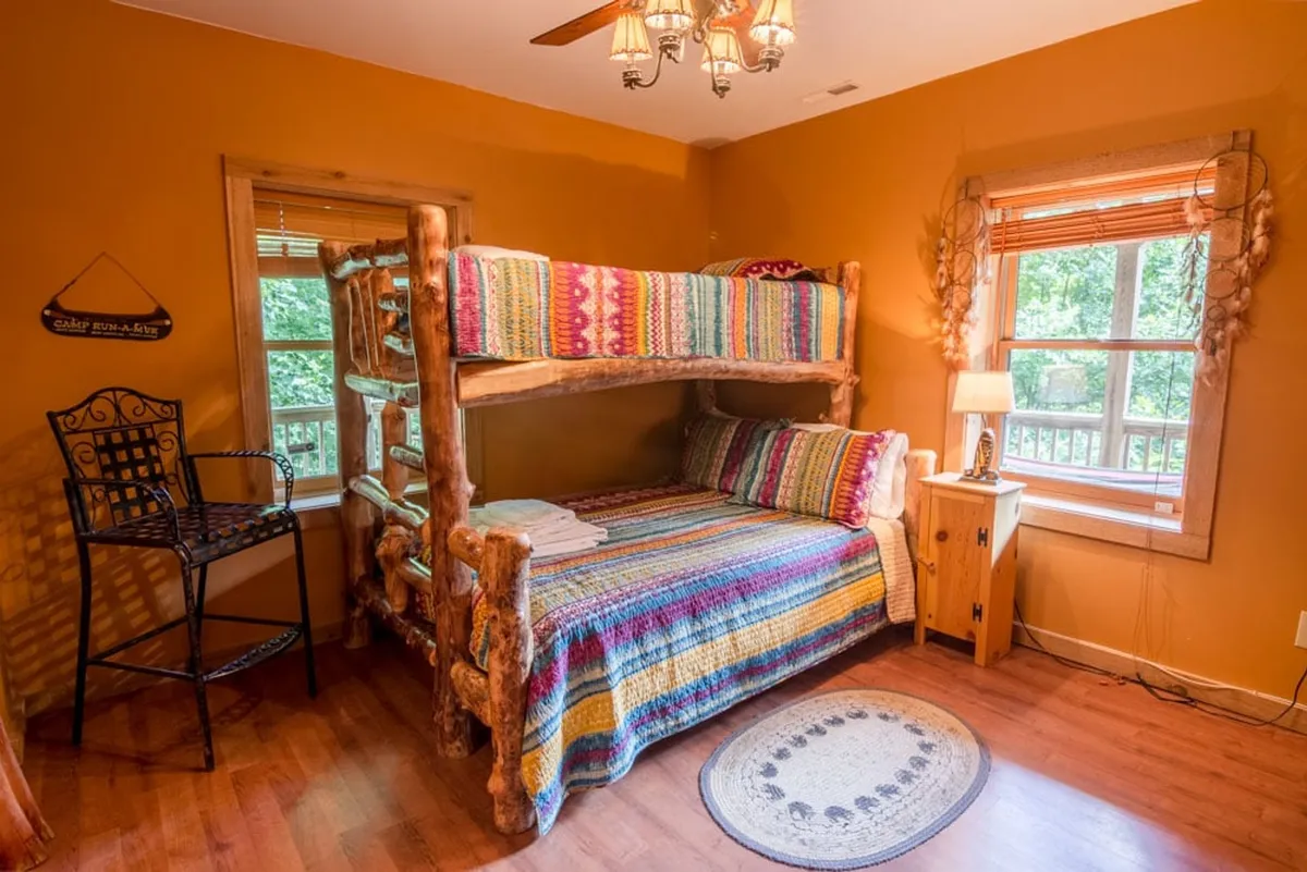 A cozy bunk room with a twin-over-queen bunk bed, colorful blankets, a nightstand with a lamp, and a decorative chair. The space is cheerful yet private, ideal for children, teens, or extra guests, and is filled with morning light.