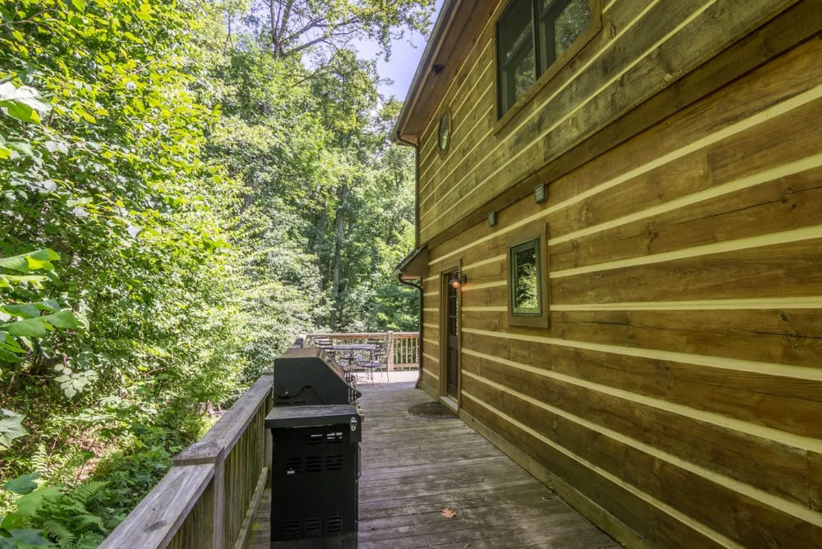 An expansive wooden deck features a high-end gas grill and plenty of deck space for outdoor activities. The area is bordered by lush greenery, hinting at the secluded and peaceful mountain setting ideal for barbecues and al fresco dining.