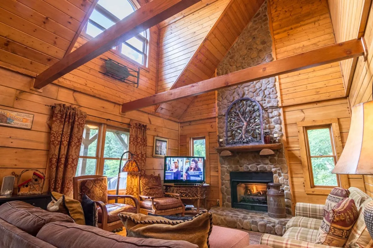 This image captures a spacious, sunlit living area featuring a prominent stone fireplace, exposed wooden beams above, large windows flooding the space with natural light, plush seating centered around the hearth, and a television visible in the background. The inviting atmosphere suggests an ideal spot for cozy evenings, socializing, and movie nights.