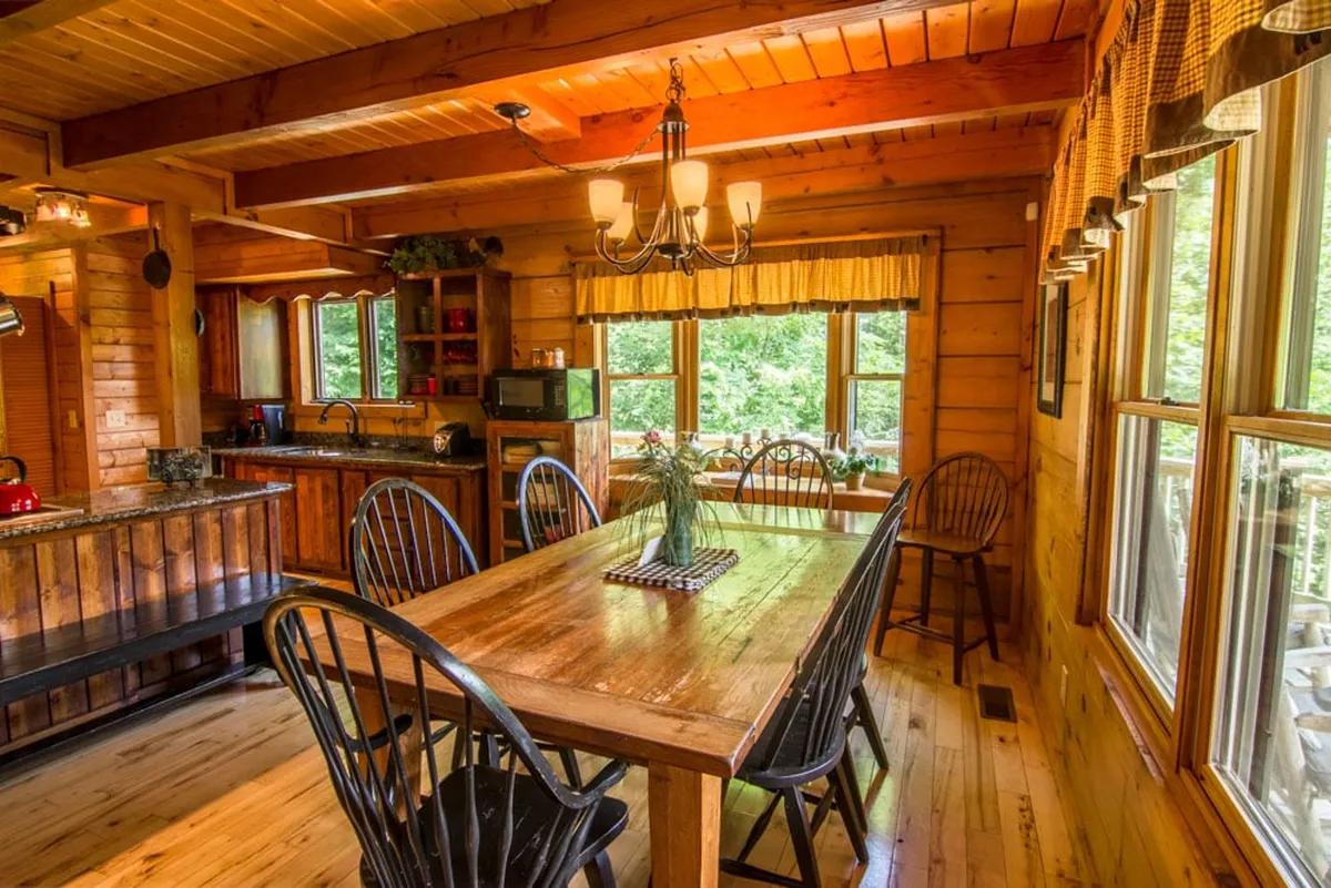 A large wooden dining table sits under a charming chandelier, adjacent to an open kitchen with custom cabinetry. Natural light streams through sizable windows, and the setting is prepared for a festive gathering, with enough space to seat a large group for meals or game nights.