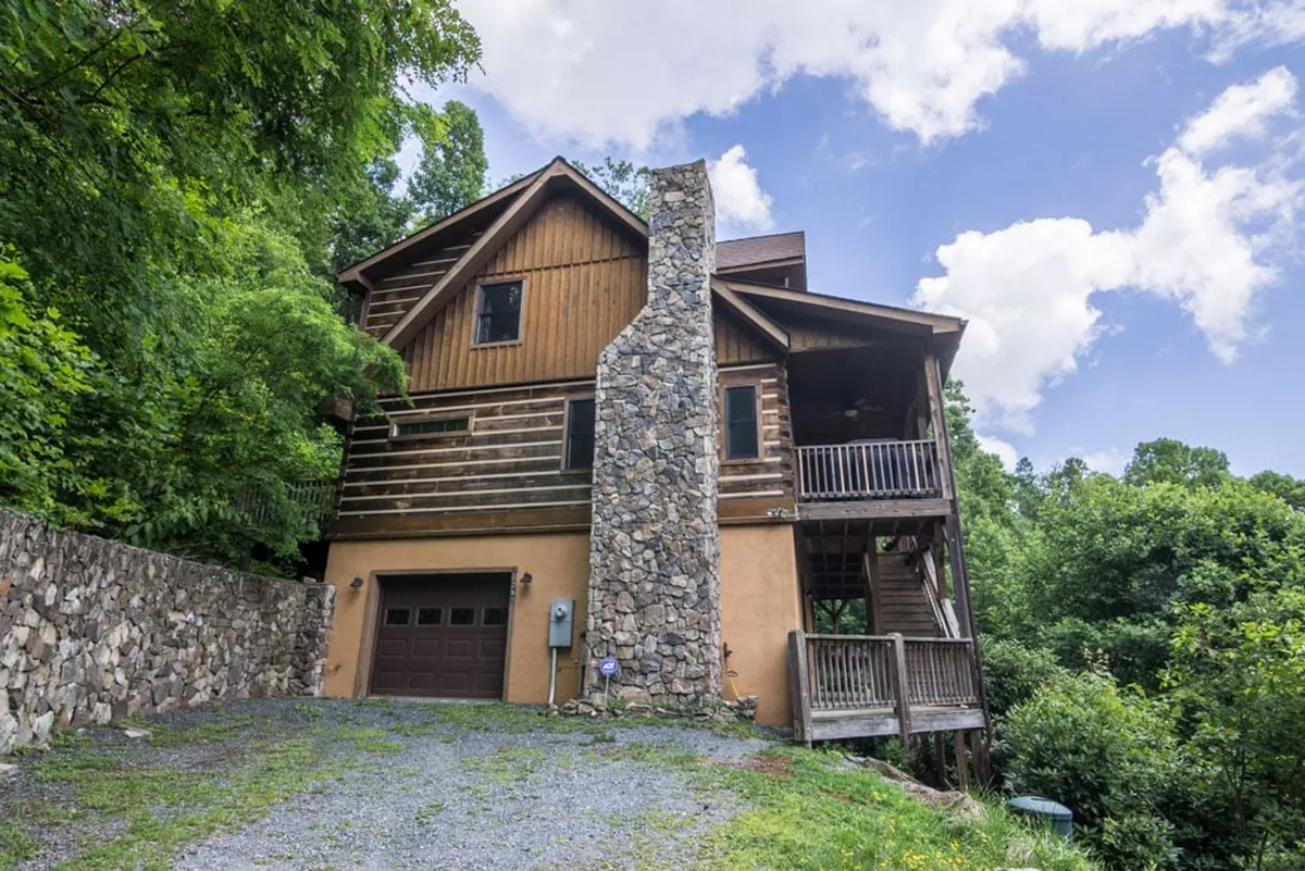 This exterior shot highlights the cabin's rustic stone chimney, wooden siding, and an inviting balcony, all surrounded by tranquil greenery. The image hints at the cabin's architectural charm and its close connection to the natural surroundings.