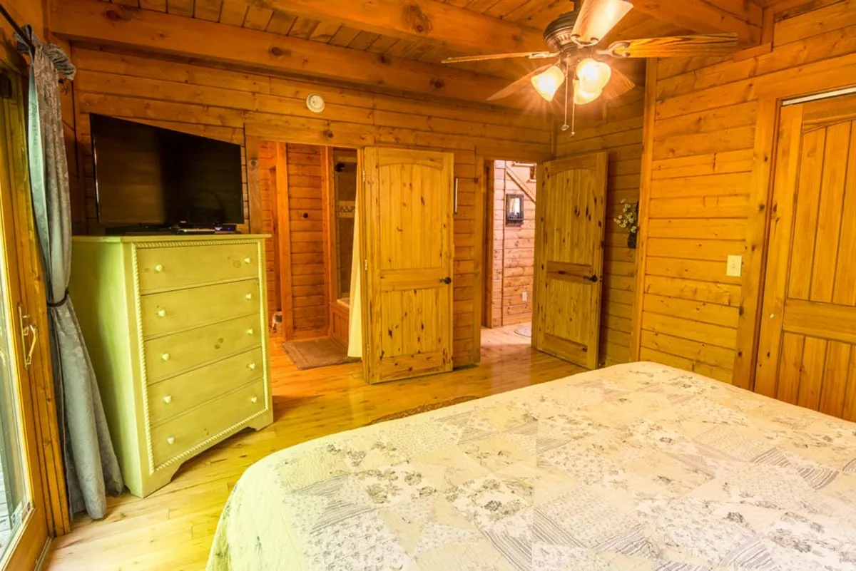A tranquil bedroom featuring wooden walls, a bed with patterned bedding, a dresser topped with a television, and open doors leading to adjoining rooms. Soft, natural light fills the space, highlighting the cozy and inviting atmosphere perfect for relaxation or late-night TV.