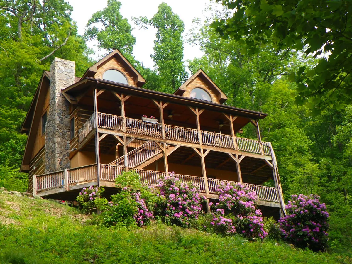 The image showcases the inviting exterior of a classic log cabin, with a welcoming wooden deck, a striking stone chimney, and bright flowering bushes. The serene setting hints at sunset views from the porch and the tranquil feeling of being surrounded by nature, perfect for relaxing or gathering with family and friends.